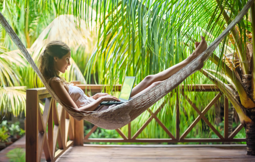 Frau arbeitet auf der Veranda am Laptop während sie in einer Hängematte liegt, umgeben von Palmen.