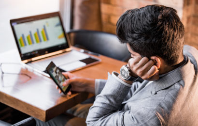 Mann mit Smartphone in der Hand prokrastiniert vor Arbeitslaptop