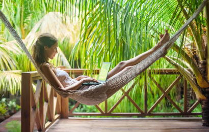 Frau arbeitet auf der Veranda am Laptop während sie in einer Hängematte liegt, umgeben von Palmen.
