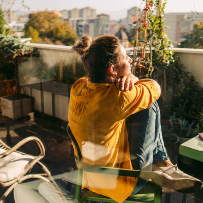 Eine junge Frau sitzt auf dem Balkon, die Beine aufgestellt und den Blick nach vorne gerichtet.