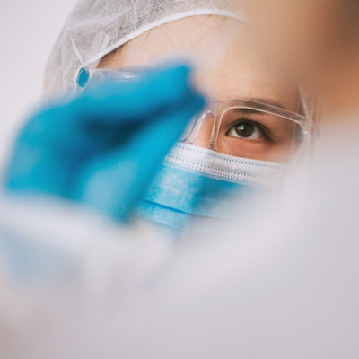 Fachkraft in Schutzkleidung mit Maske und Handschuhen nimmt mit Stäbchen Testprobe eines Patienten