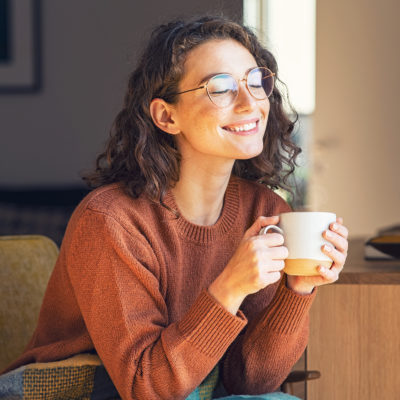 Frau sitzt lächelnd auf Couch, hält eine Tasse mit Warmgetränk  und lächelt in die Sonne