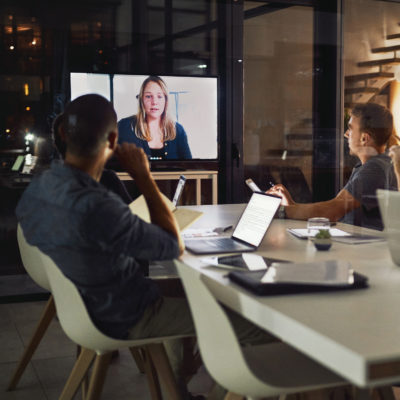 Nächtliches hybrides Meeting im Büro, in das eine Kollegin per Video dazugeschaltet ist,