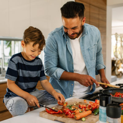 Junger Mann und Kind stehen in der Küche, schneiden und braten Gemüse und  haben Spaß dabei