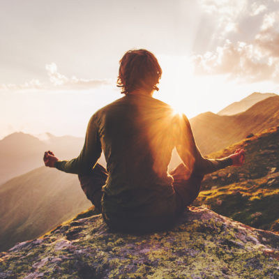 Mann sitzt in meditierender Pose auf einem Berg und blickt in den Sonnenuntergang