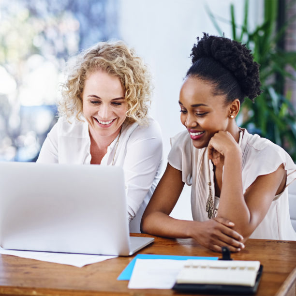 Zwei Frauen sitzen im Büro gemeinsam am Laptop und lachen bei der Arbeit