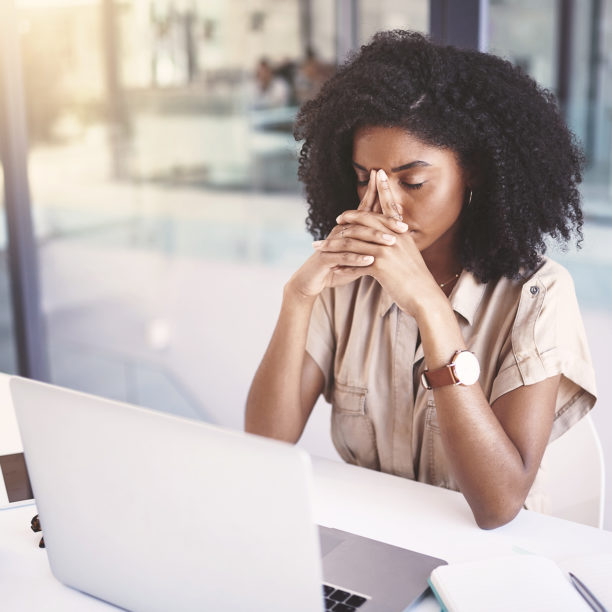 MEDISinn Angebot Burnout Prävention Junge Frau sitzt am Schreibtisch im Büro, hat die Augen geschlossen und hält Hände vors Gesicht