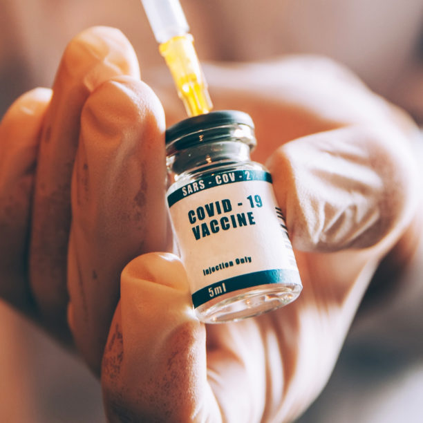 Hand einer Fachkraft greift mit Handschuhen COVID-Impfflasche und zieht mit Ampulle eine Dosis