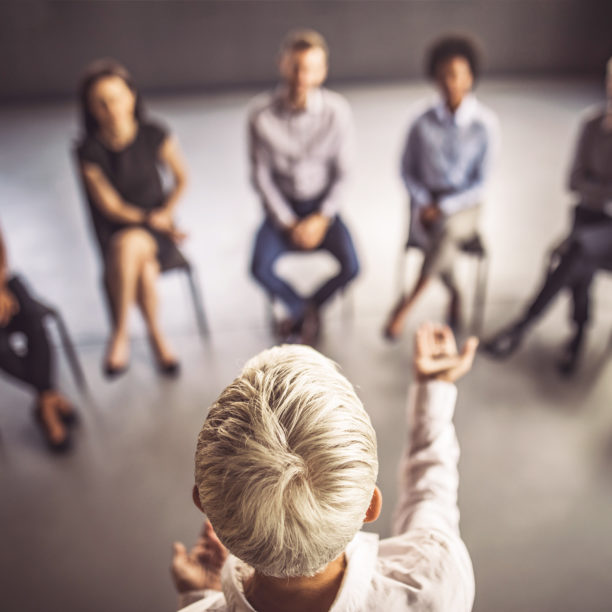 MEDISinn Angebot Führungskräftecoaching Frau spricht und gestikuliert vor einer Gruppe Menschen, die in einem Stuhlkreis vor ihr sitzen
