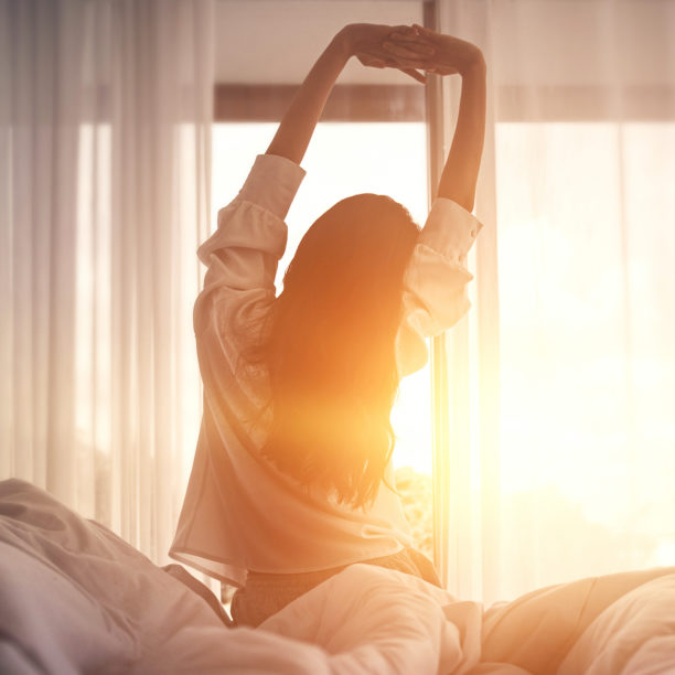 Frau richtet sich morgens im Bett auf während die Sonne aufgeht und streckt sich sitzend