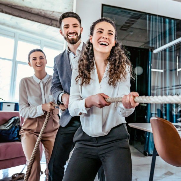 MEDISinn Angebot Konflikte im Team lösen Mehrere Kolleg:innen im Büro ziehen gemeinsam an einem Tau und lachen dabei