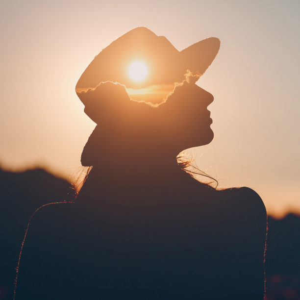 Frau mit Hut sitzt im Dunkeln während die Sonne untergeht und sich in ihrem Hut symbolisch spiegelt