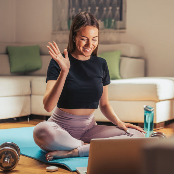 Junge Frau in Sportoutfit sitzt auf Yogamatte vor Laptop und winkt anderen virtuell zu
