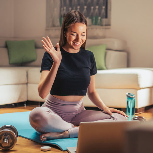 MEDISinn Angebot Online Fitnessstudio Junge Frau in Sportoutfit sitzt auf Yogamatte vor Laptop und winkt anderen virtuell zu