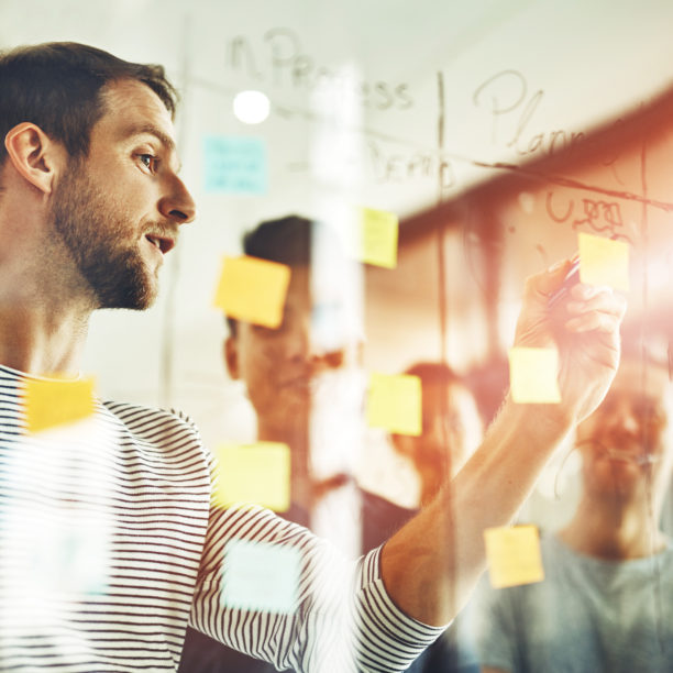 MEDISinn Angebot Systemisches Coaching Mitarbeiter:innen scribbeln Notizen auf eine Glaswand mit Stift und Post its.