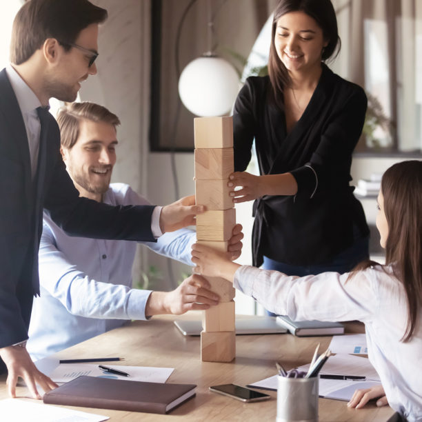 MEDISinn Angebot Teambuilding- und entwicklung Junges Team sitzt und steht an Meetingtisch und baut mit Holzsteinen gemeinsam einen Turm