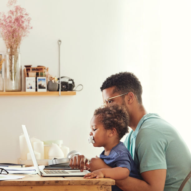 Mann und Kind sitzen am zu Hause eingerichteten Arbeitsplatz zusammen und blicken in den Laptop