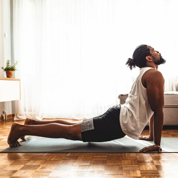 MEDISinn Angebot Yoga Junger Mann führt Yogaposition herabschauender Hund auf Yogamatte im Wohnzimmer aus