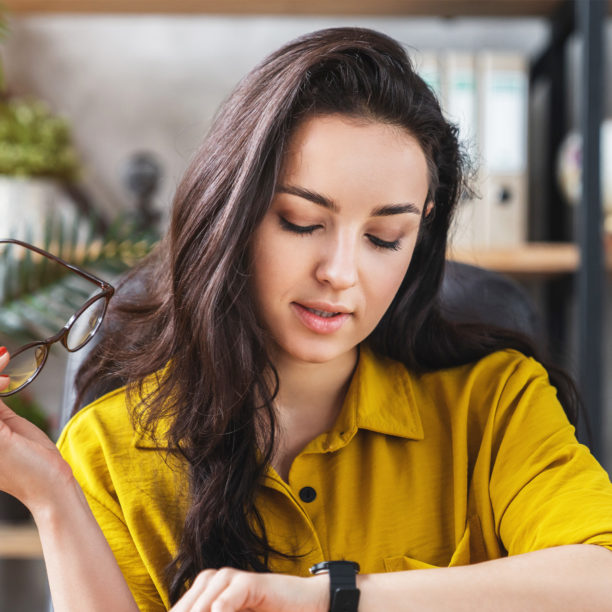 MEDISinn Angebot Zeitmanagement Junge Frau sitzt im Büro und prüft die Zeit auf ihrer Armbanduhr